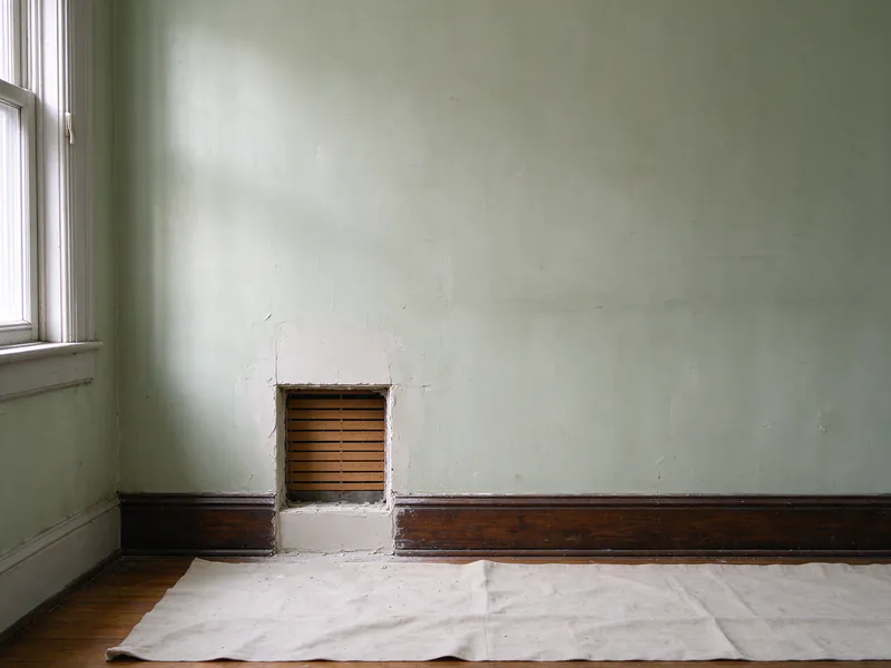 Before: square access opening showing wood lath in a pale green plaster wall above a dark baseboard, with a drop cloth on the floor