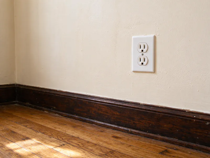 New white three-prong outlet in a cream plaster wall above an original dark-stained baseboard and oak floor