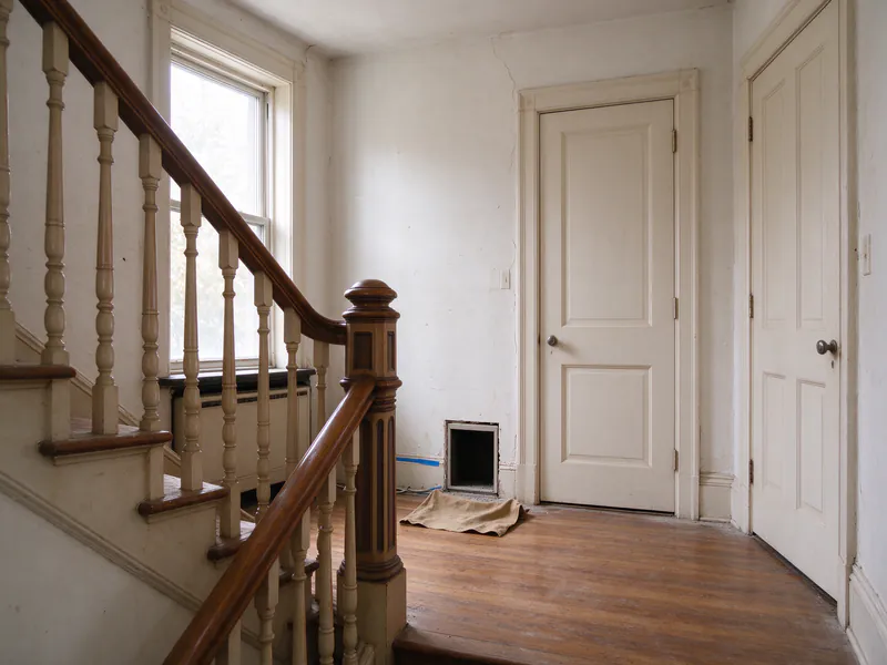 Stair landing in a converted Philadelphia rowhouse with a turned-wood banister, two unit doors and a small access opening over a drop cloth